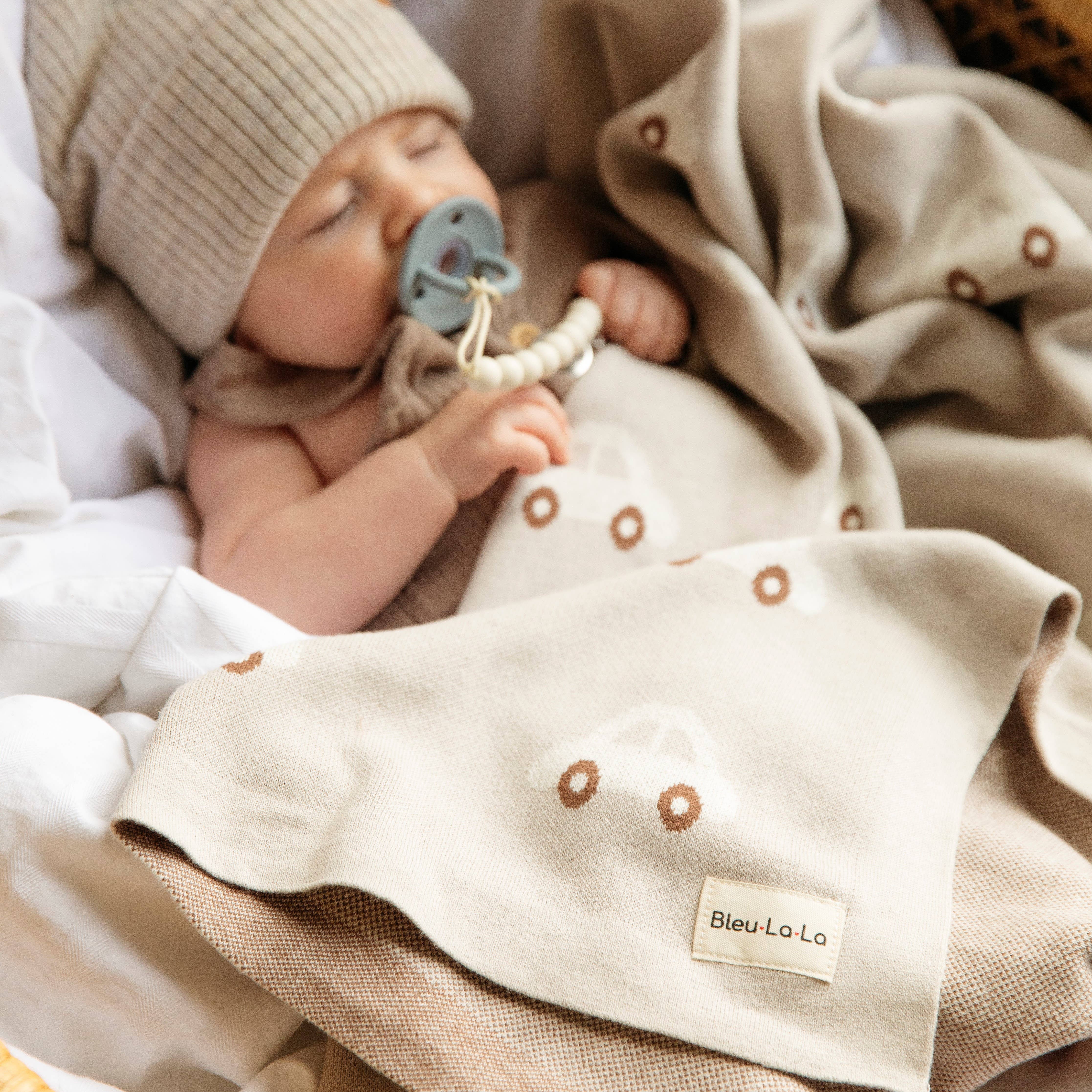 Baby wrapped in a beige blanket with a pacifier, wearing a knit hat, on a soft surface.