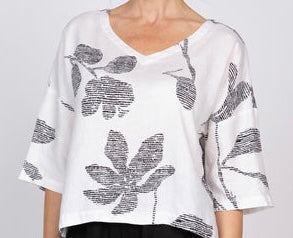 IMG_0128 (front, outdoor, dark-haired model): Woman wearing white linen Tessa top with bold black abstract floral print, v-neck and dropped shoulders, styled with printed wide-leg pants, outdoor setting
IMG_0129 (front, studio, blonde model): Front view of white linen Tessa top with oversized black abstract floral print and v-neckline, styled with black wide-leg pants on neutral background
IMG_0132 (full-length, outdoor, dark-haired model): Full-length front view of white linen Tessa top with bold black flo