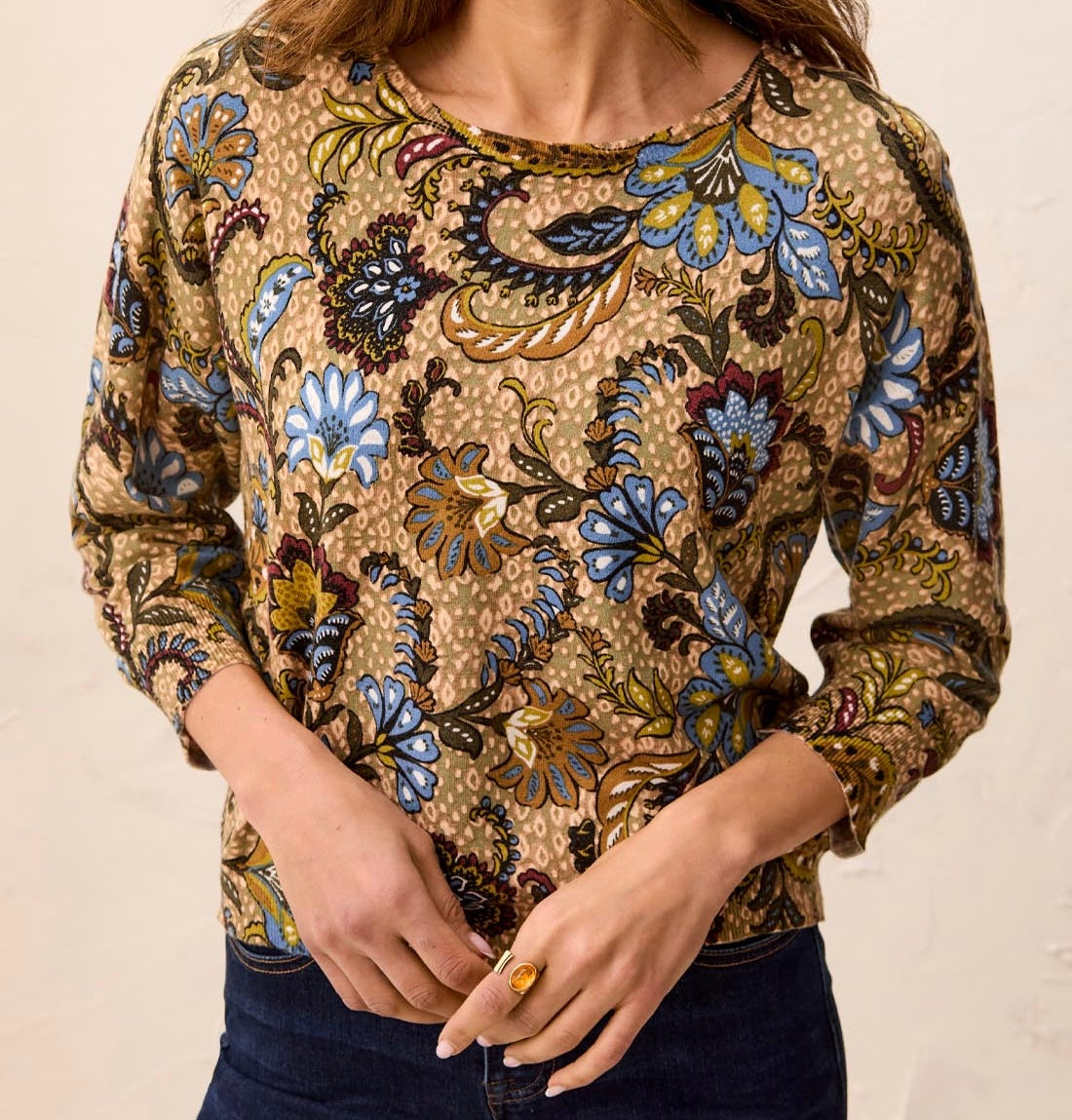 A woman wearing a beige long-sleeve top with a colorful paisley and floral print in blue, brown, and gold, paired with dark blue jeans.
