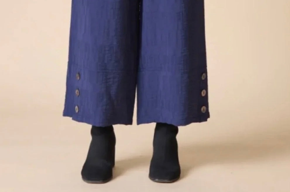 A close-up of a person's lower legs wearing wide-leg blue trousers with decorative buttons on the cuffs and black ankle boots, set against a plain beige studio background.