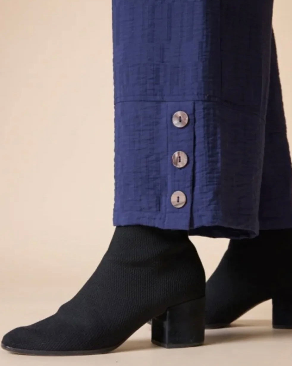 Close-up of a person wearing dark navy ankle boots with a block heel and wide-leg blue trousers with three silver buttons at the hem, set against a plain beige background.
