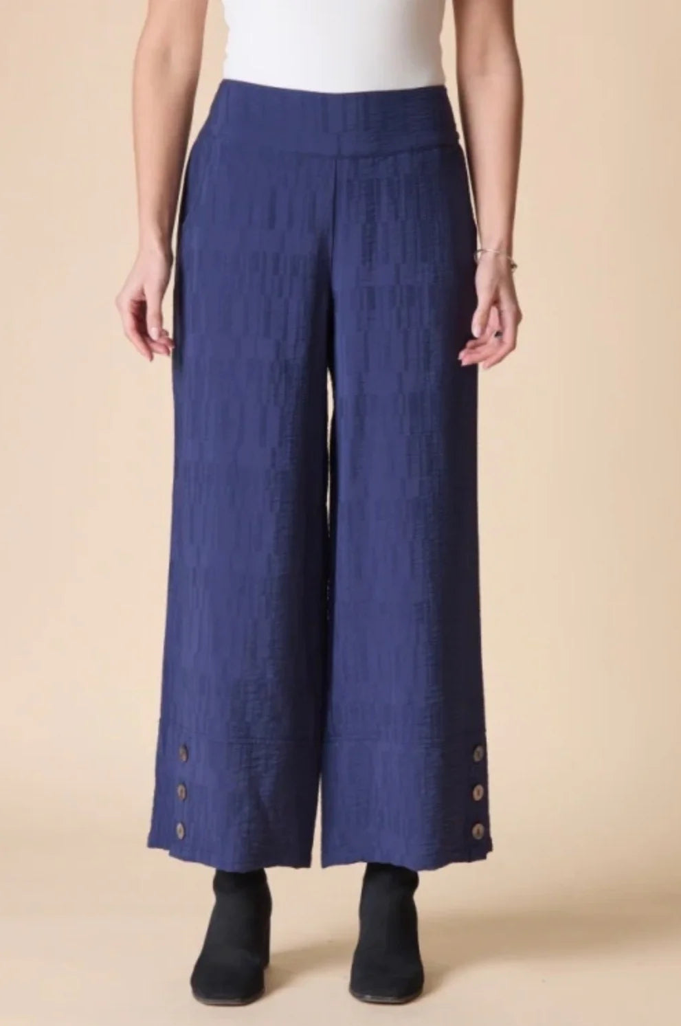 A model is shown from the waist down wearing high-waisted, wide-leg cropped pants in a textured navy blue fabric, styled with a white top and black ankle boots against a solid beige background.