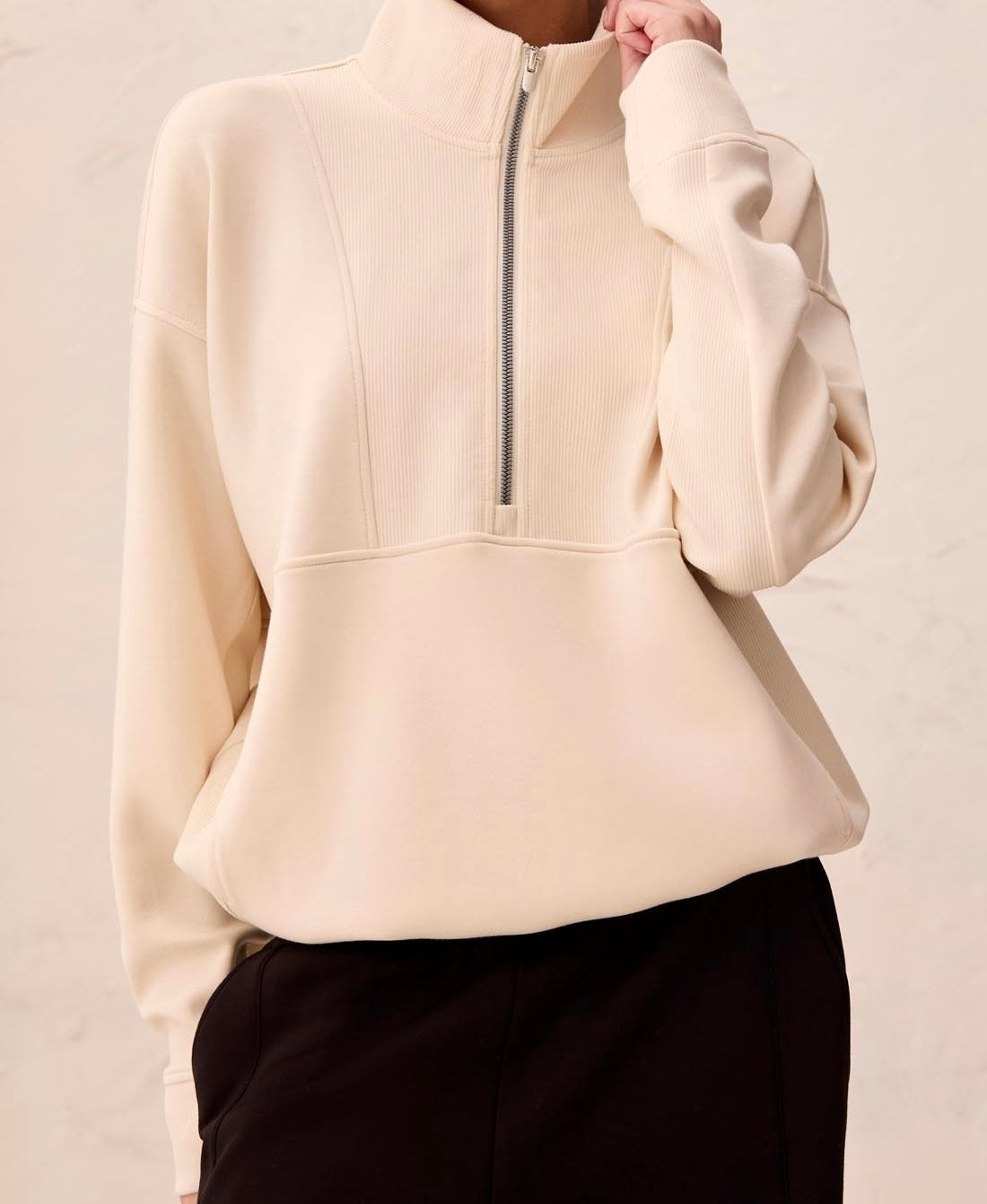 A person wearing a cream-colored ribbed half-zip pullover sweatshirt with a stand collar and black pants, shown from the neck down against a light textured wall.