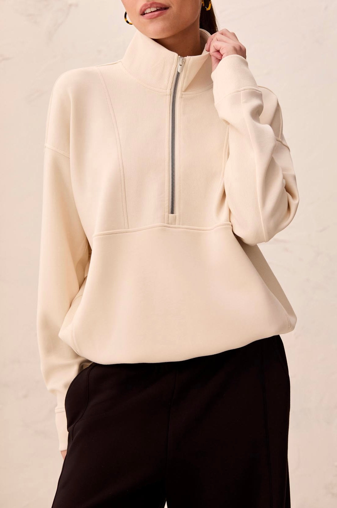 A woman wearing a cream-colored quarter-zip pullover sweatshirt with a mock neck and black pants, standing against a light textured wall.
