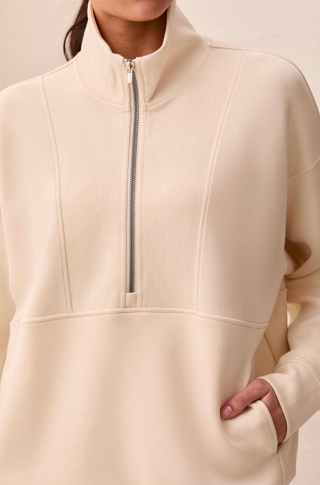 A woman shown from the back wearing a cream-colored mock-neck knit sweater and dark wide-leg trousers, standing against a textured off-white wall.