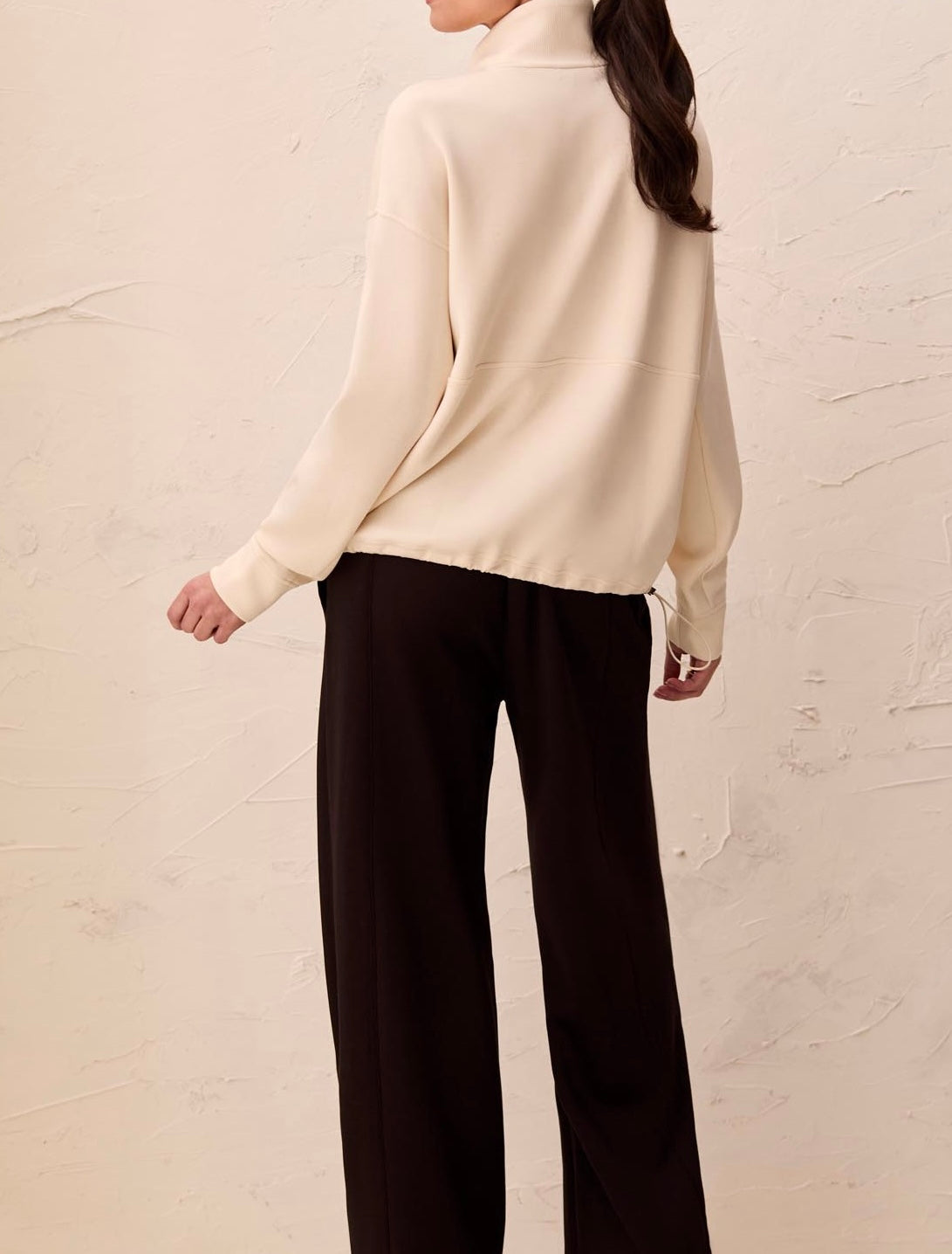 A woman shown from the back wearing a cream-colored mock-neck knit sweater and dark wide-leg trousers, standing against a textured off-white wall.