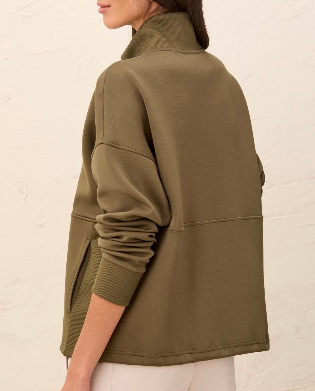 A woman wearing an olive green fleece pullover jacket, shown from a side-back angle against a textured off-white wall.