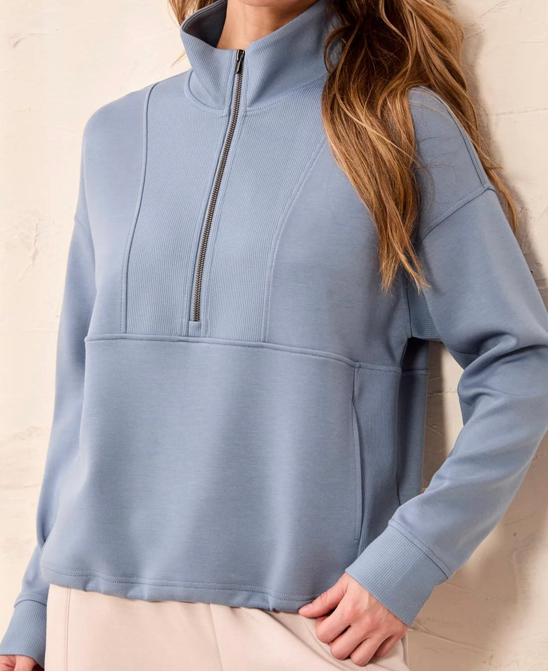A woman wearing a light blue quarter-zip pullover sweatshirt with a mock neck and ribbed front panels, paired with light beige pants, standing against a textured off-white wall.