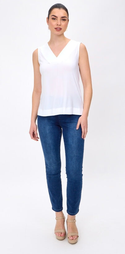 A woman modeling a white sleeveless V-neck top paired with dark blue skinny jeans and beige heeled sandals, standing against a plain white studio background.