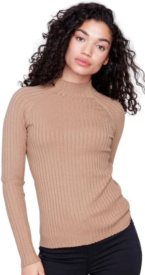 A woman with long, curly dark hair wearing a beige ribbed mock neck sweater and black pants, photographed against a plain white background.