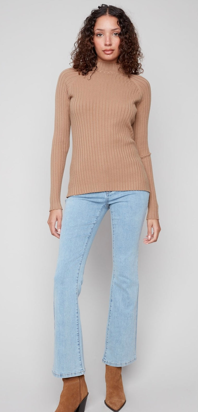 A woman modeling a beige ribbed mock-neck long-sleeve sweater paired with light-wash blue jeans and brown ankle boots, shot against a neutral light gray background.