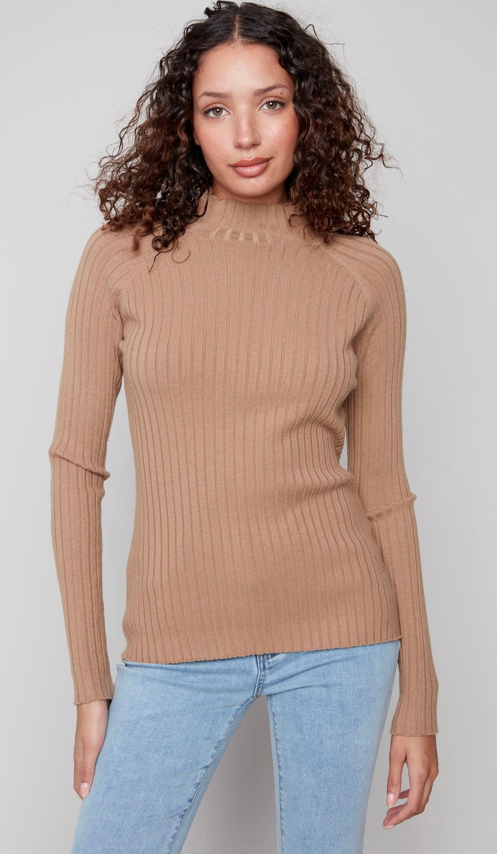 A woman with curly brown hair wearing a ribbed knit mock neck sweater in camel beige and light blue jeans, standing against a light gray studio background.
