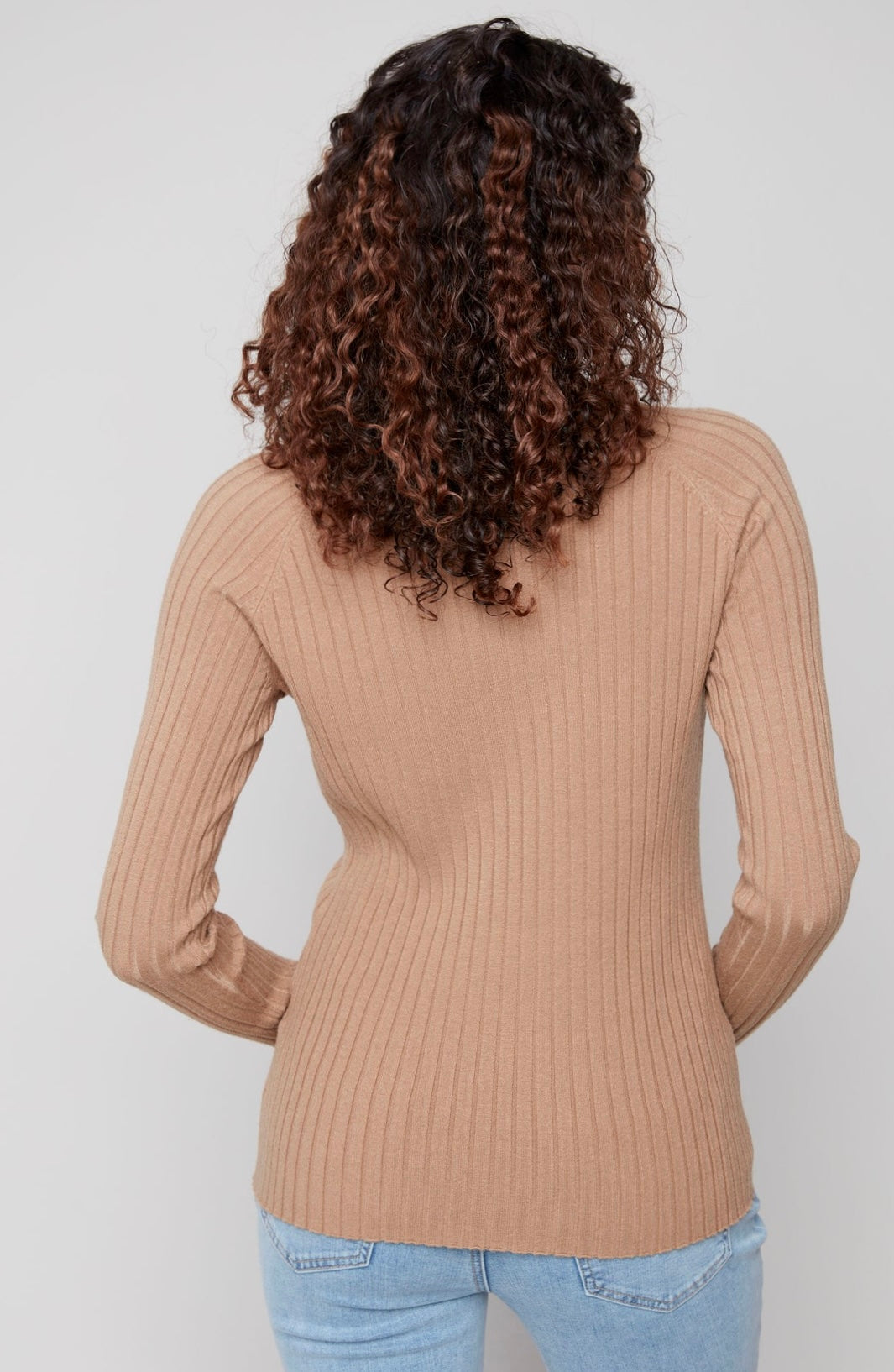 A woman viewed from the back wearing a beige ribbed long-sleeve knit top and light blue jeans against a plain light gray studio background.