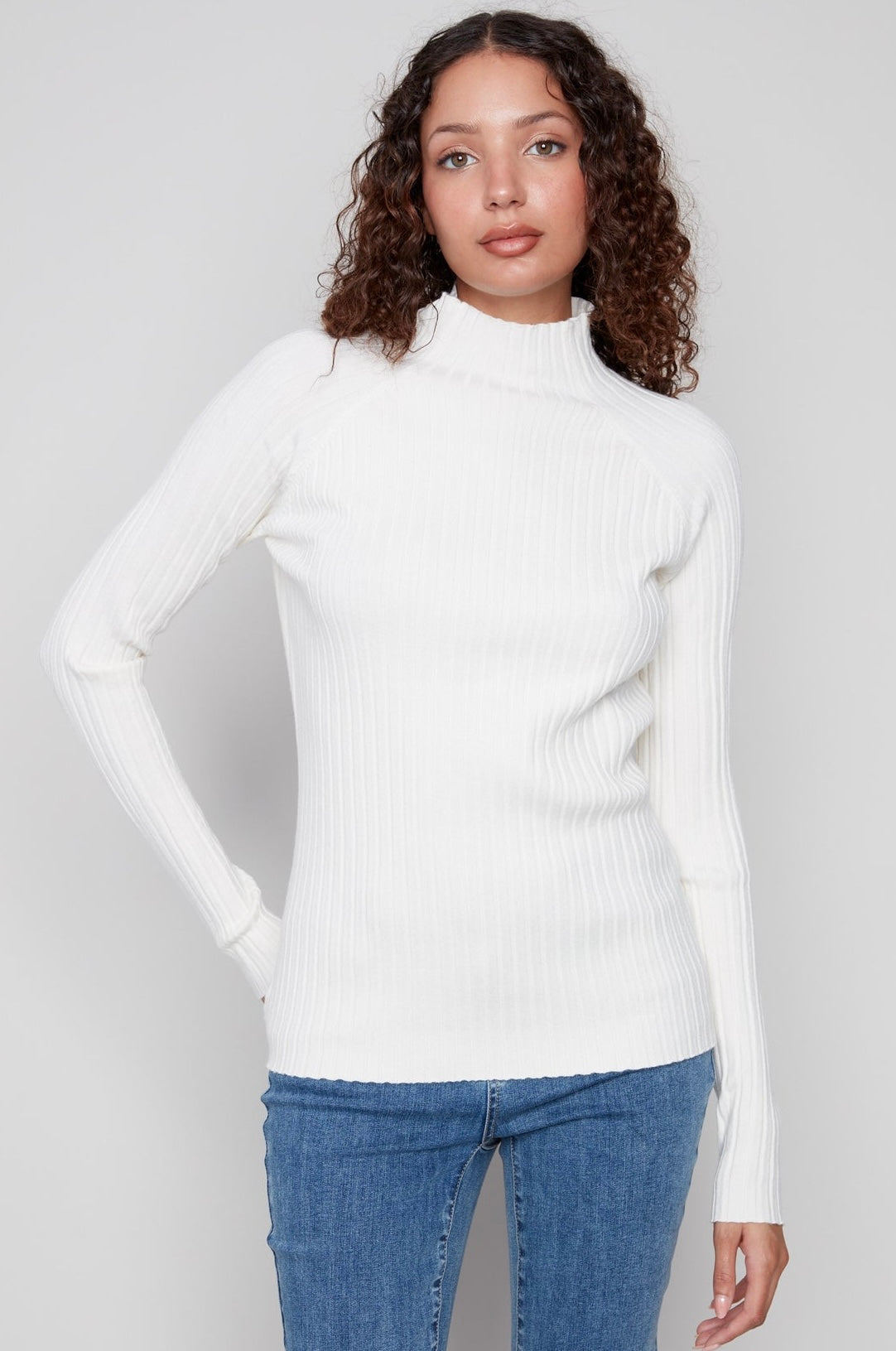 A woman wearing a white ribbed mock neck long-sleeve sweater and blue jeans, shown from the neck down against a light gray background.