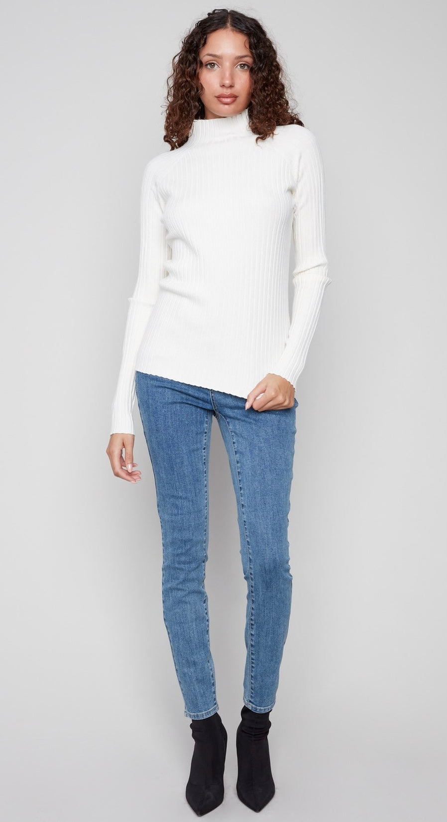 A woman modeling a white ribbed mock neck long-sleeve sweater paired with blue skinny jeans and black ankle boots, standing against a light gray studio background.