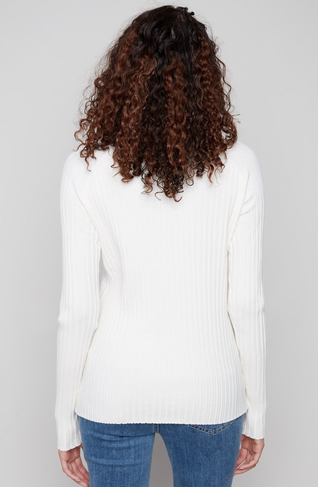 A woman viewed from the back wearing a white ribbed long-sleeve sweater and blue jeans against a light gray studio background.