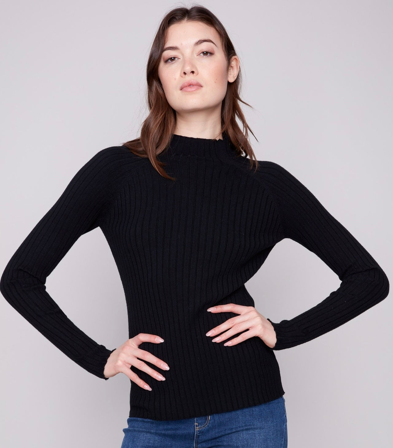 A woman modeling a black ribbed mock neck sweater with long sleeves, paired with blue jeans, against a solid light gray background.