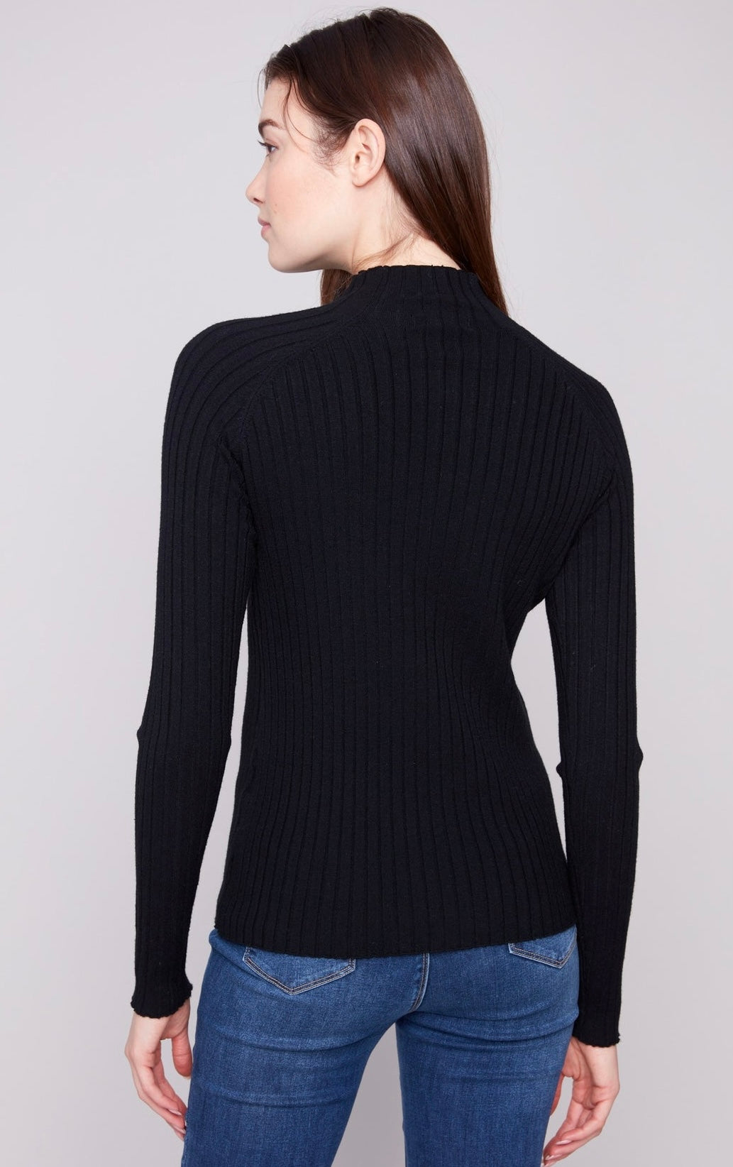 A woman shown from the back wearing a black ribbed mock neck long-sleeve sweater and blue jeans against a light gray studio background.