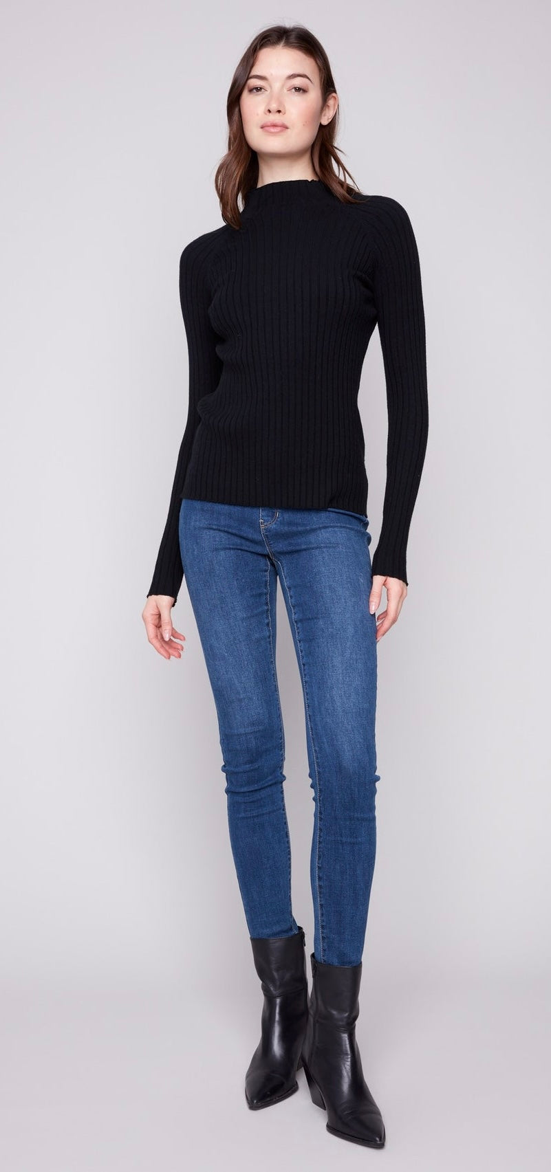 A woman modeling a black ribbed mock neck sweater, blue skinny jeans, and black ankle boots against a light gray studio background.