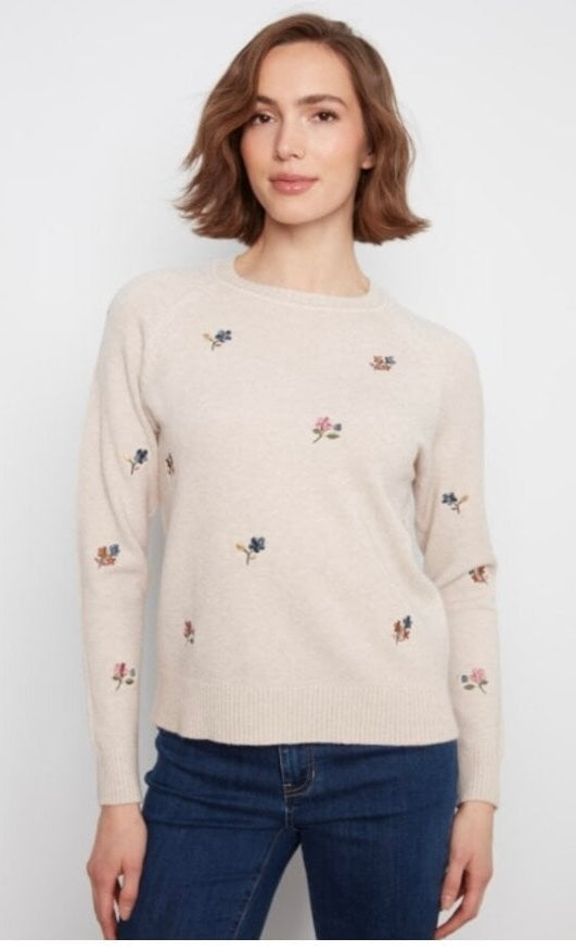 A woman wearing a light beige crewneck sweater with small embroidered floral details, paired with dark blue jeans, against a plain light gray background.