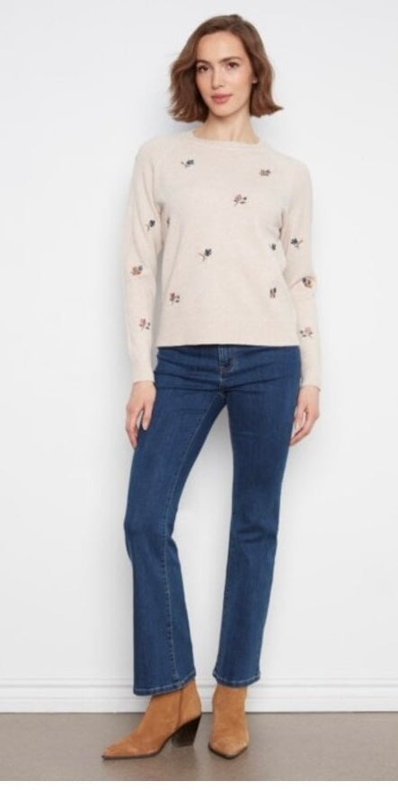 A woman modeling a light beige crewneck sweater with small embroidered details, paired with dark blue flare jeans and tan ankle boots, standing against a white wall.