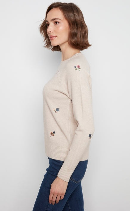 A woman wearing a cream-colored knit sweater with small embroidered floral motifs, paired with dark blue jeans, shown from a side profile against a light gray background.