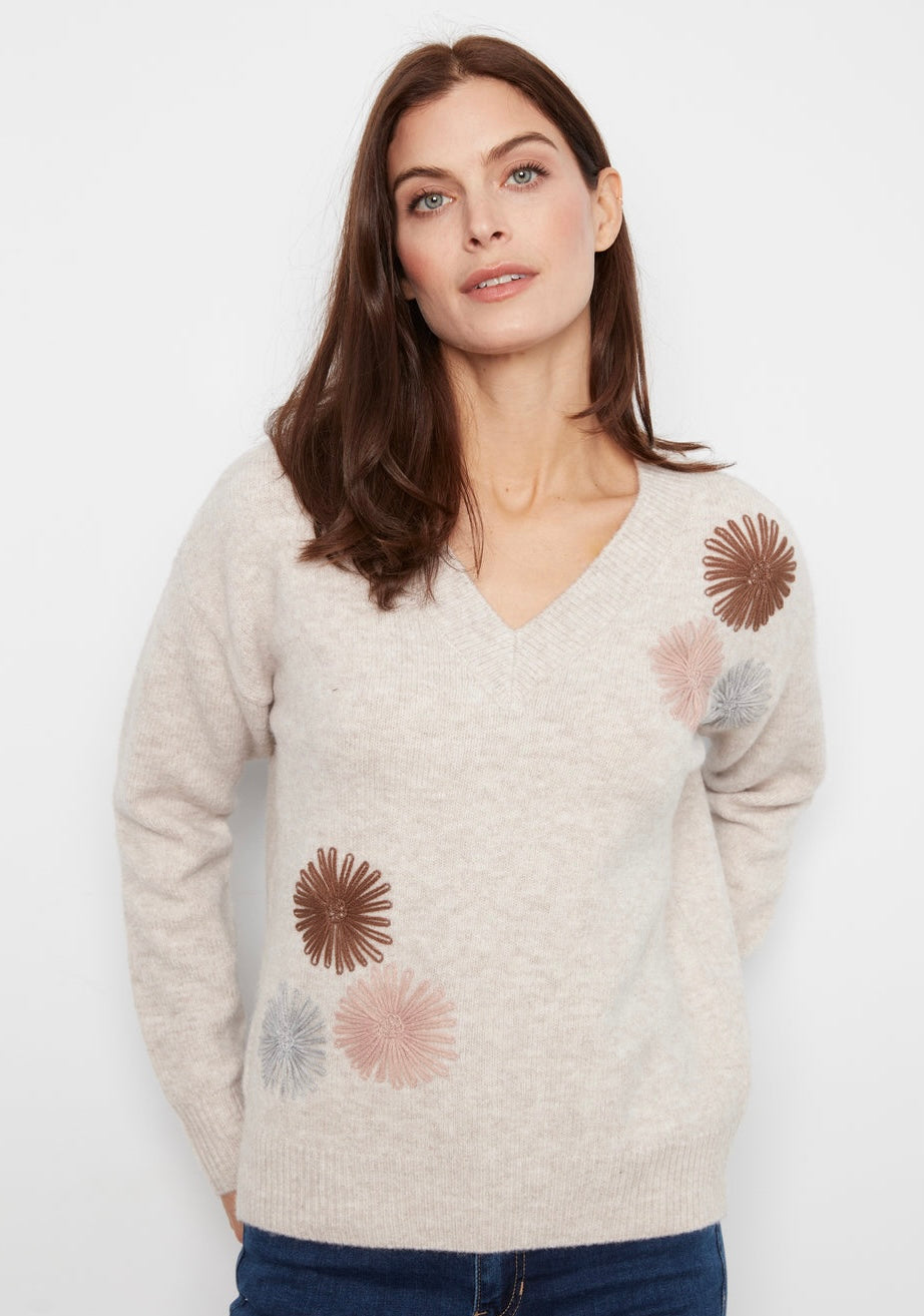 A woman stands against a white wall wearing a cream knit sweater with brown and pink floral patches, dark blue skinny jeans, and black ankle boots.