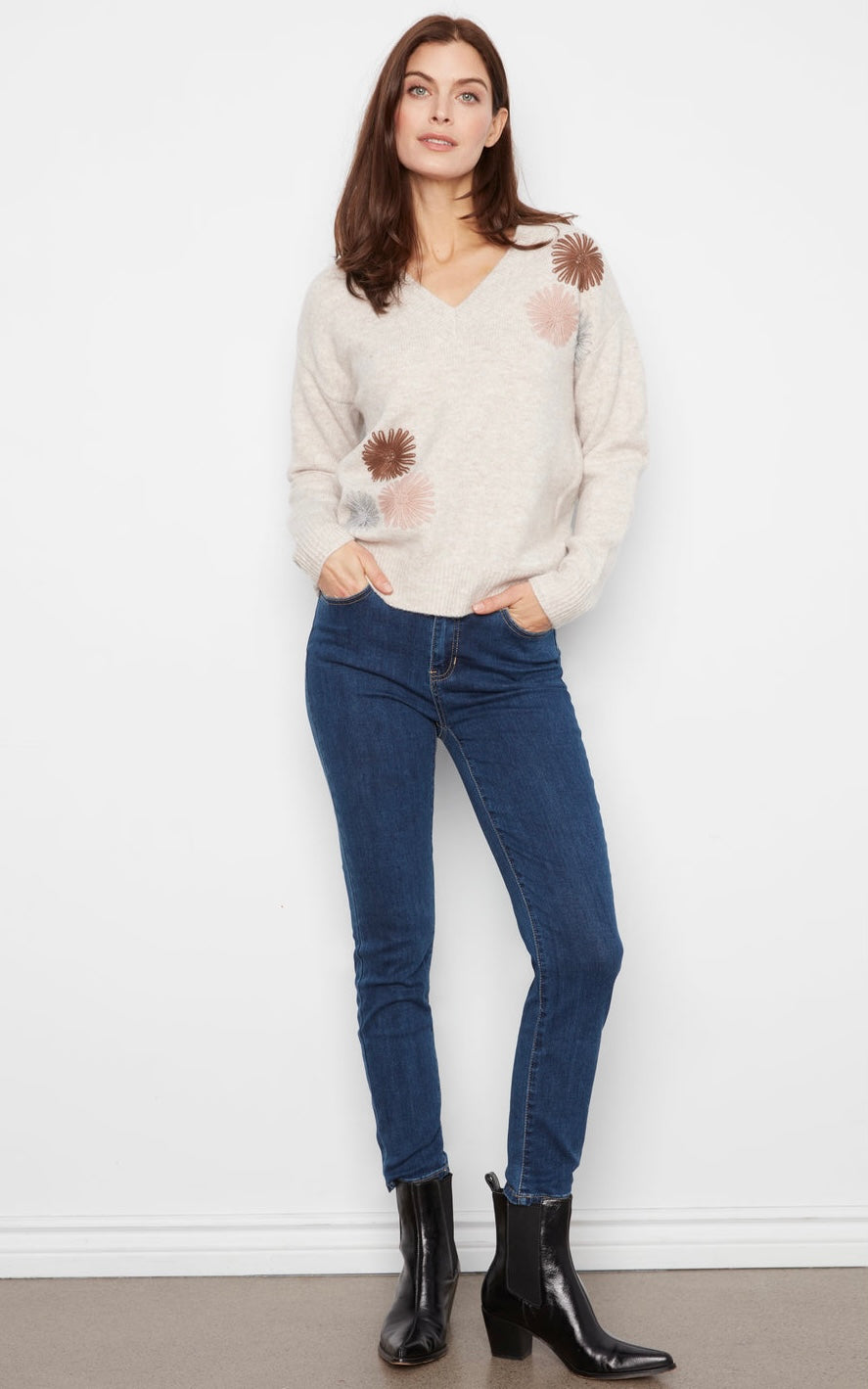 A woman stands against a white wall wearing a cream knit sweater with brown and pink floral patches, dark blue skinny jeans, and black ankle boots.
