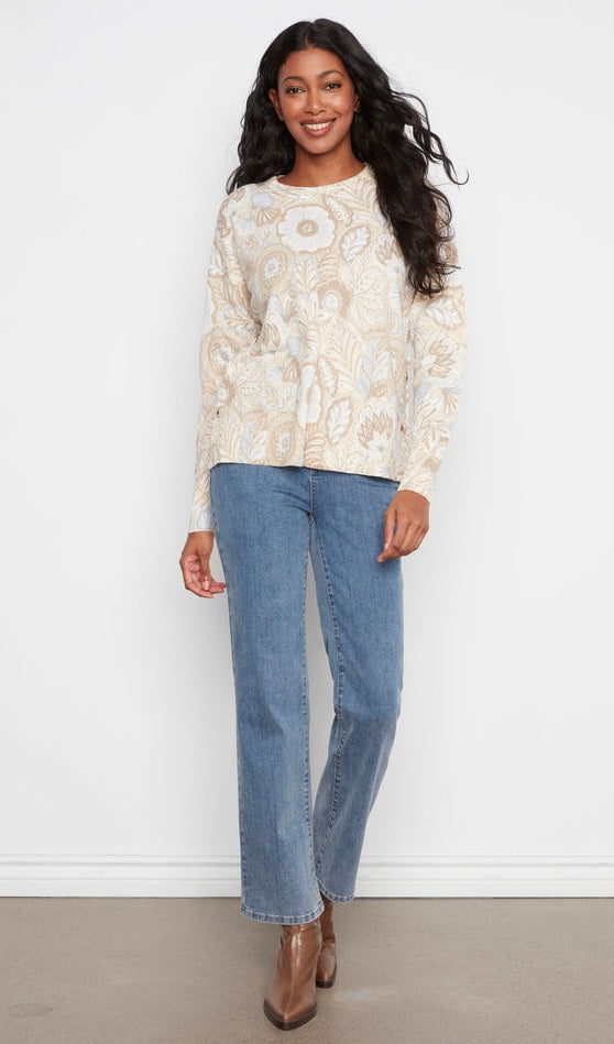 A woman modeling a cream and beige paisley patterned knit sweater paired with blue jeans and brown ankle boots, standing against a white wall.
