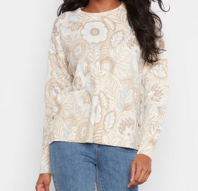 A woman wearing a cream and beige floral knit sweater with blue jeans, shown from the shoulders down against a plain white background.