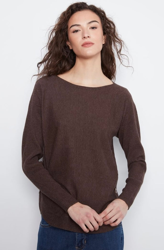 A woman wearing a dark brown boat-neck knit sweater and blue jeans, standing against a plain light gray background.