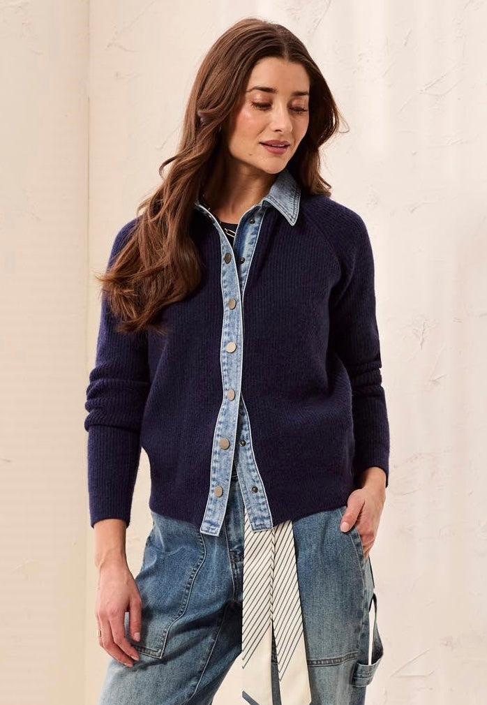 A woman wearing a navy blue ribbed knit cardigan over a light blue denim shirt, paired with blue jeans and a striped tie, standing against a light beige textured wall.