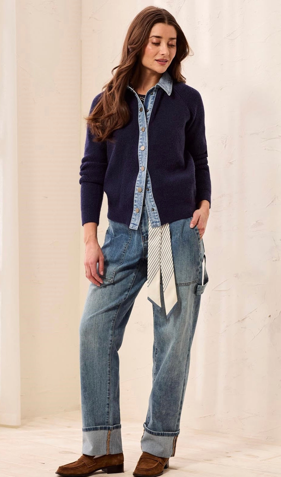 A woman modeling a navy blue knit cardigan over a light blue denim shirt, paired with light-wash blue jeans, a striped tie, and brown leather shoes, set against a neutral cream-colored wall.