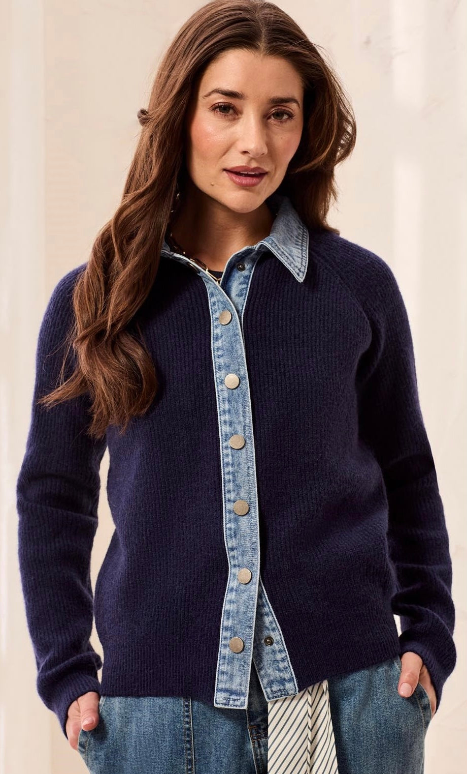 A woman wearing a navy blue ribbed knit cardigan with a light blue denim button-front placket and collar, layered over a matching denim shirt and paired with jeans.