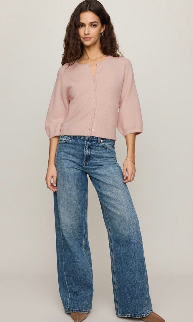 A woman modeling a light pink three-quarter sleeve button-up top with a keyhole neckline, paired with wide-leg blue jeans, against a plain light gray studio background.