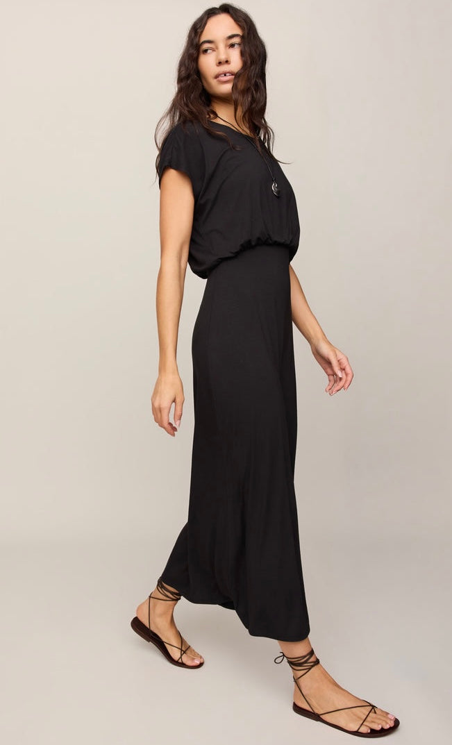 A woman modeling a black short-sleeve maxi dress with a gathered waist and side buttons, paired with black lace-up flat sandals, against a light gray studio background.