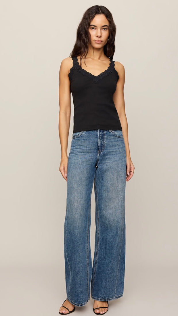 A woman modeling a black sleeveless camisole top with delicate scalloped trim and wide-leg blue denim jeans, standing against a solid light beige studio background.