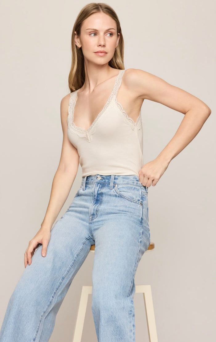 A woman wearing a beige lace-trimmed camisole top and light-wash blue jeans, posed against a plain light beige studio background.