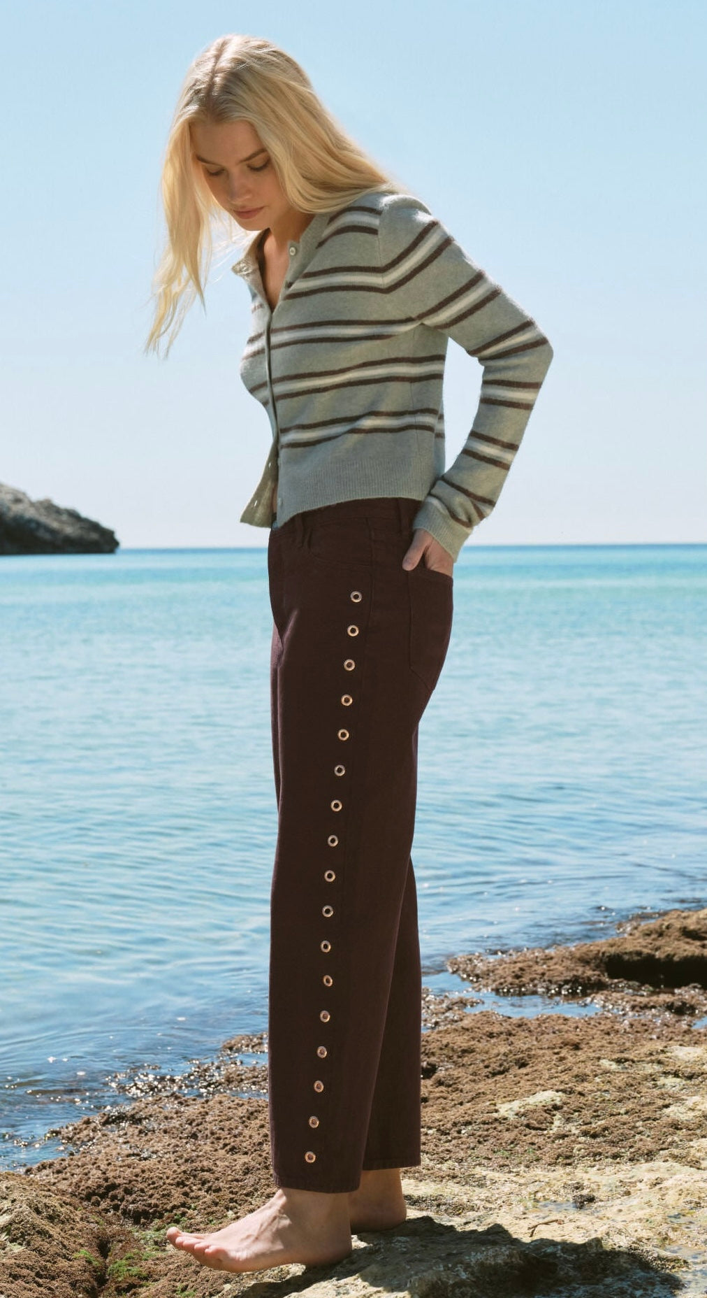 A woman stands barefoot on a rocky shoreline with the ocean and blue sky behind her, wearing a striped knit top and dark brown wide-leg pants with a vertical row of gold-tone buttons down the side.