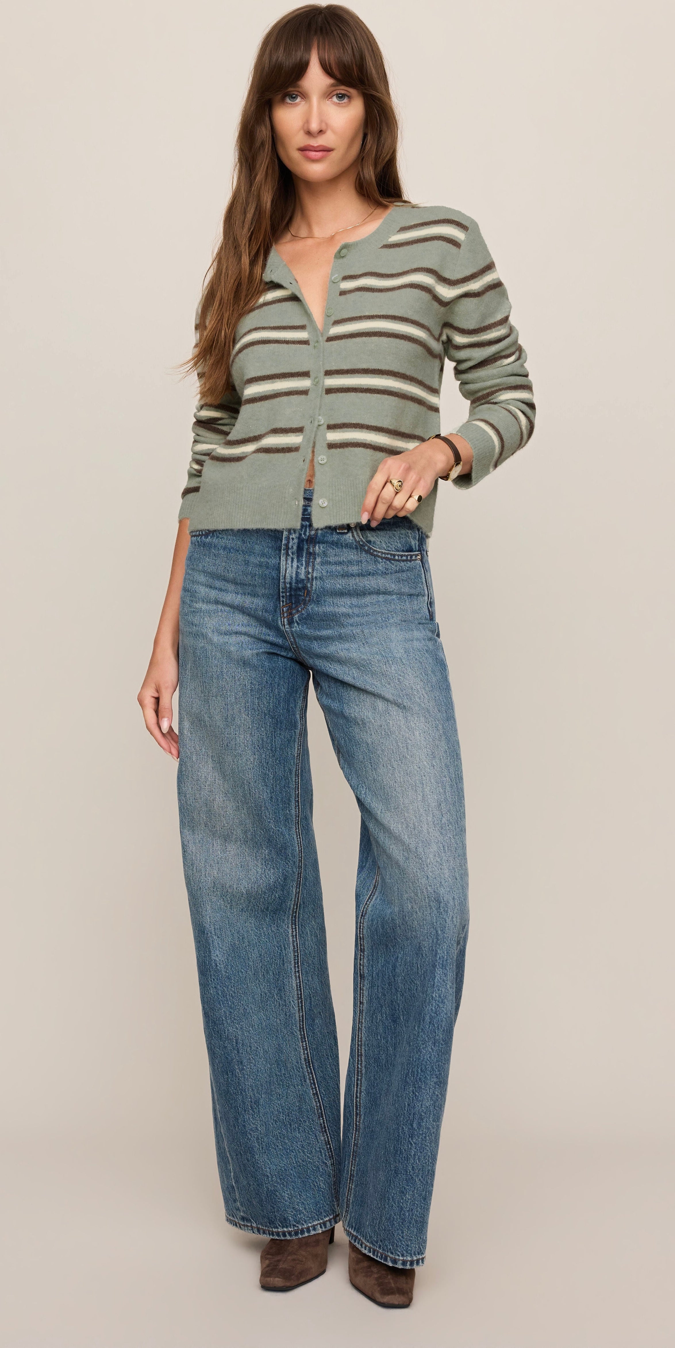 A woman modeling a cropped, long-sleeve knit cardigan with horizontal olive green, beige, and brown stripes, paired with wide-leg blue jeans and brown boots, against a solid light beige studio background.