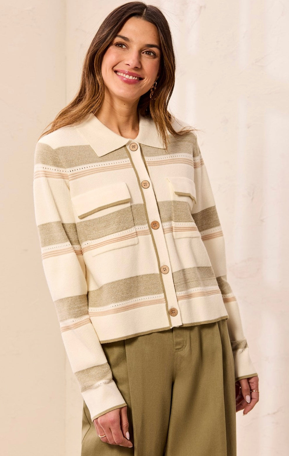 A woman wearing a cream and beige striped knit button-up cardigan with a collar and brown buttons, paired with olive green pants, set against a neutral beige background.