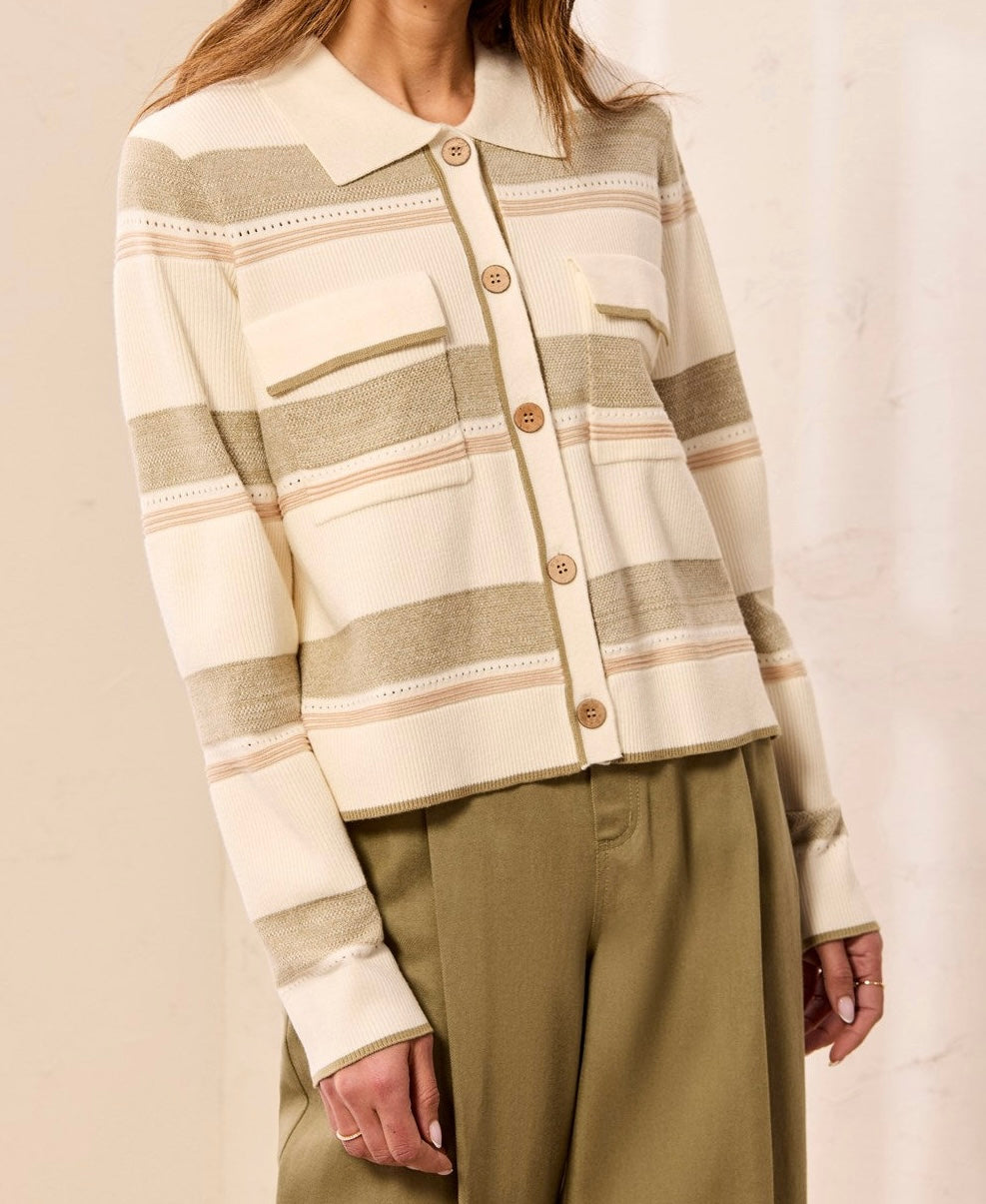 A woman wearing a cream and beige striped knit cardigan with brown buttons, paired with olive green pants, against a soft beige background.
