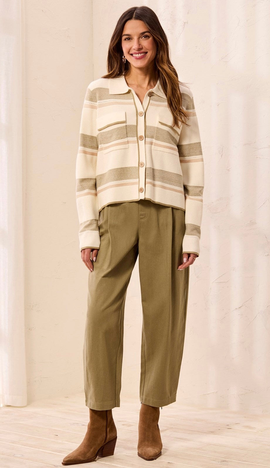 A woman modeling a cream and tan striped button-up knit cardigan with olive green cropped trousers and brown ankle boots, standing against a light beige curtain backdrop.