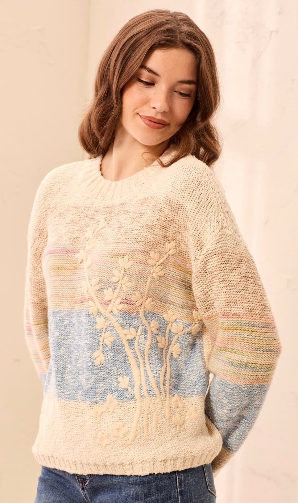 A woman wearing a cream-colored knit sweater with a colorful horizontal stripe pattern in blue, pink, and yellow, featuring a raised floral motif on the front.