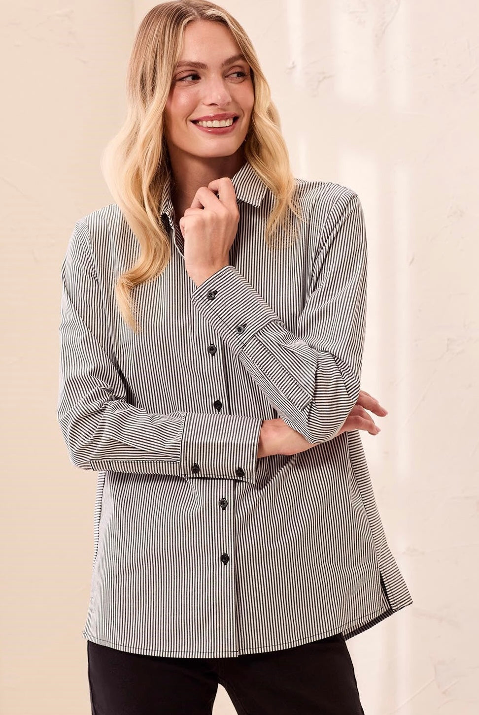 A woman modeling a light gray long-sleeve button-up shirt with black pants and black loafers, standing in a brightly lit indoor setting with a cream-colored wall and light wood floor.