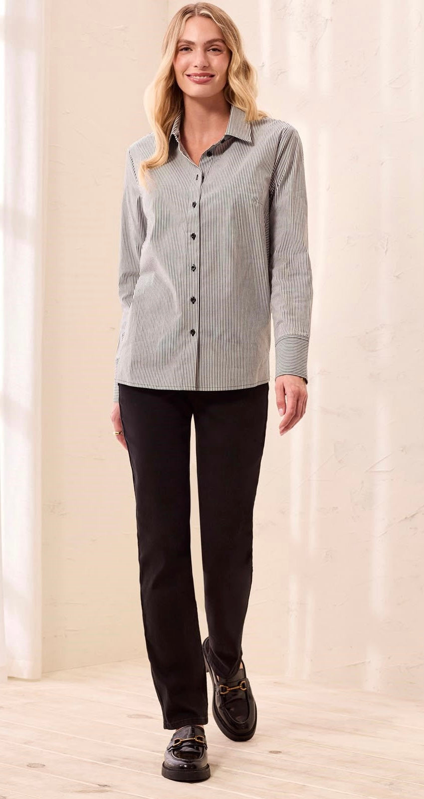 A woman modeling a light gray long-sleeve button-up shirt with black pants and black loafers, standing in a brightly lit indoor setting with a cream-colored wall and light wood floor.