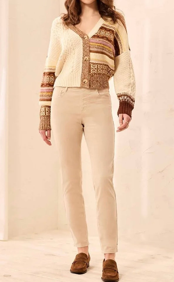 A woman wearing a cream and brown patterned knit cardigan, beige pants, and brown suede loafers, standing against a light beige background.