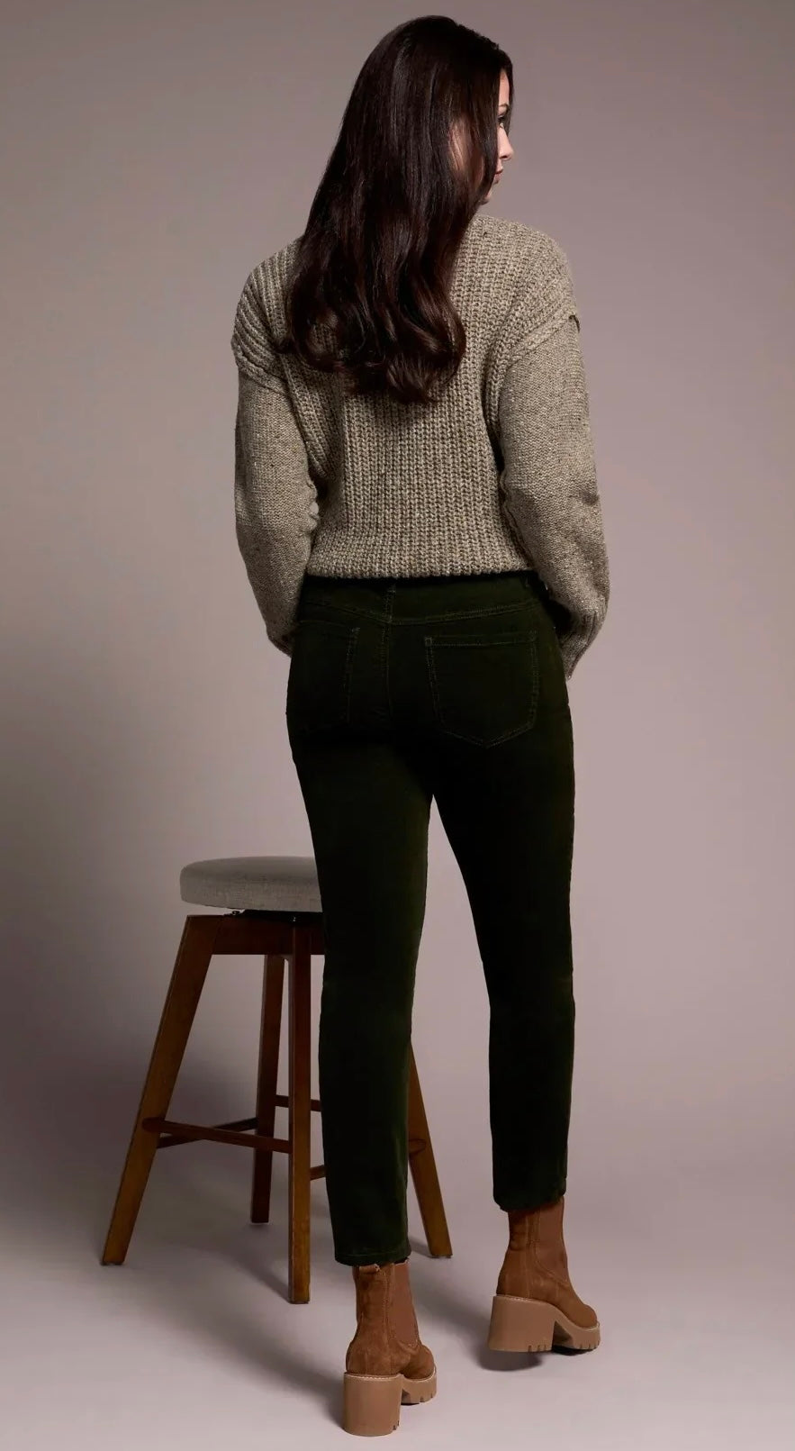 A woman is shown from behind wearing a textured light gray knit sweater, dark olive green skinny jeans, and brown ankle boots, standing next to a wooden stool against a neutral studio background.