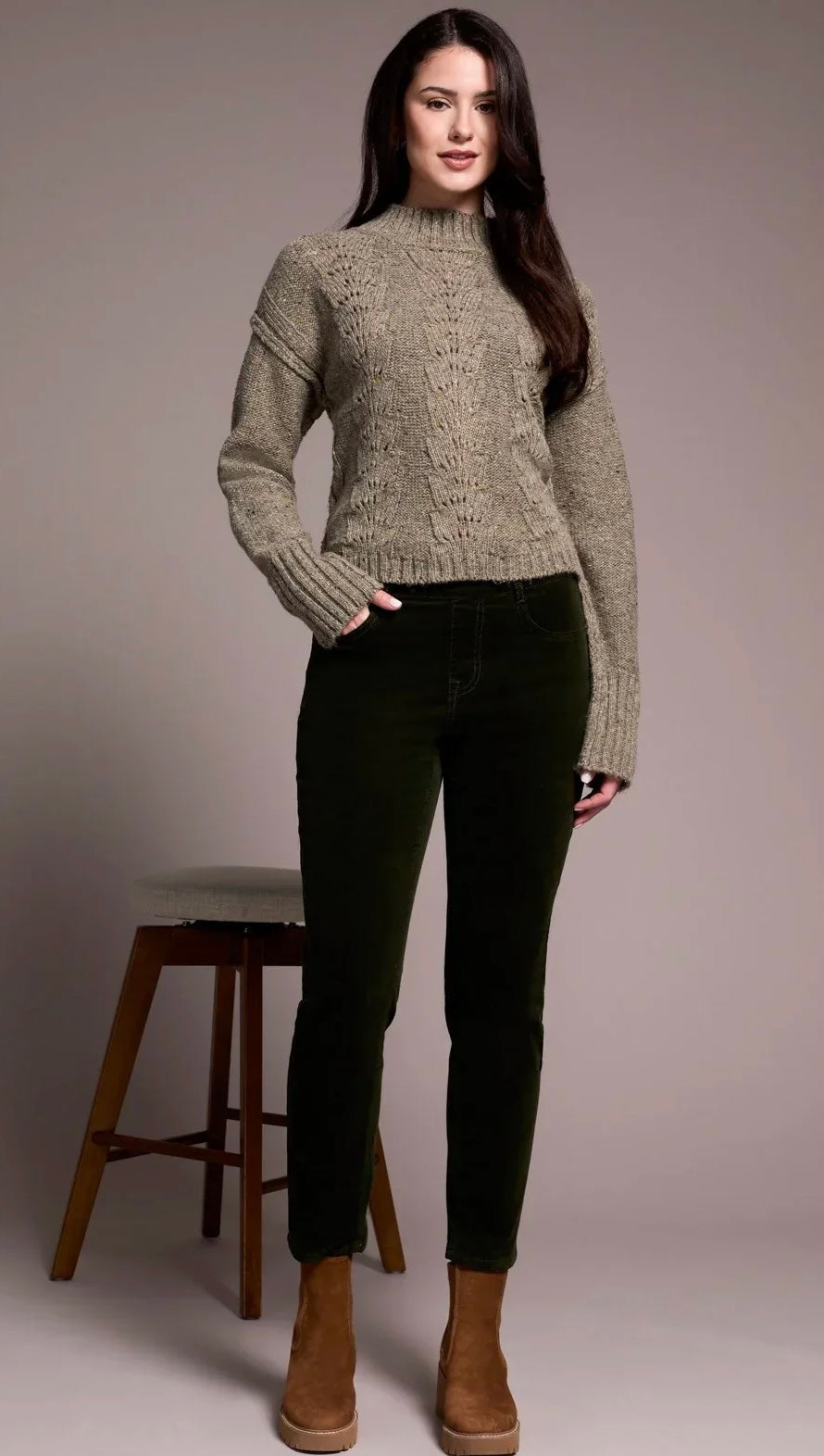 A woman modeling a beige cable-knit mock-neck sweater paired with dark green skinny pants and brown suede ankle boots, standing next to a wooden stool against a neutral studio backdrop.