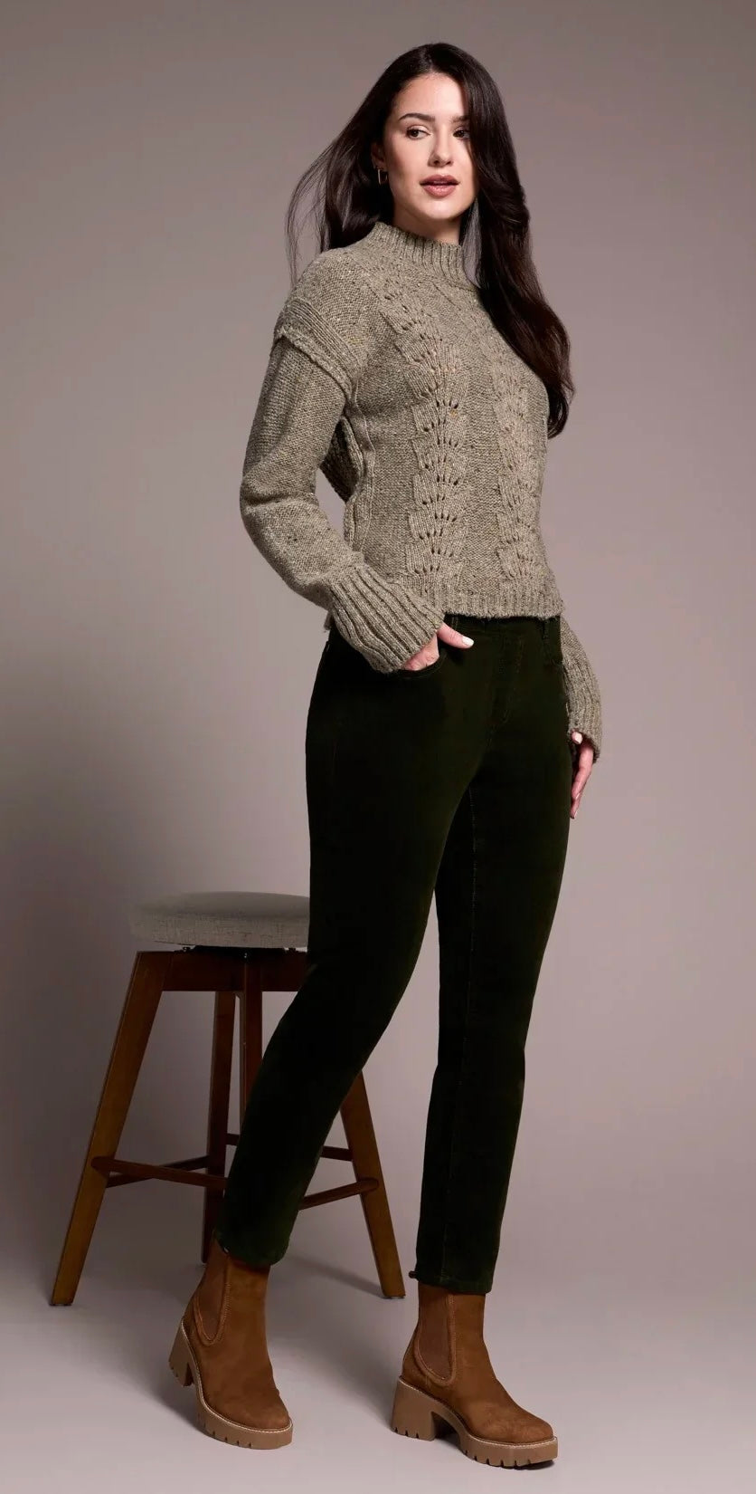 A woman modeling a textured knit mock-neck sweater in a warm gray-beige color, paired with dark green pants and brown ankle boots, standing next to a wooden stool against a solid gray studio background.